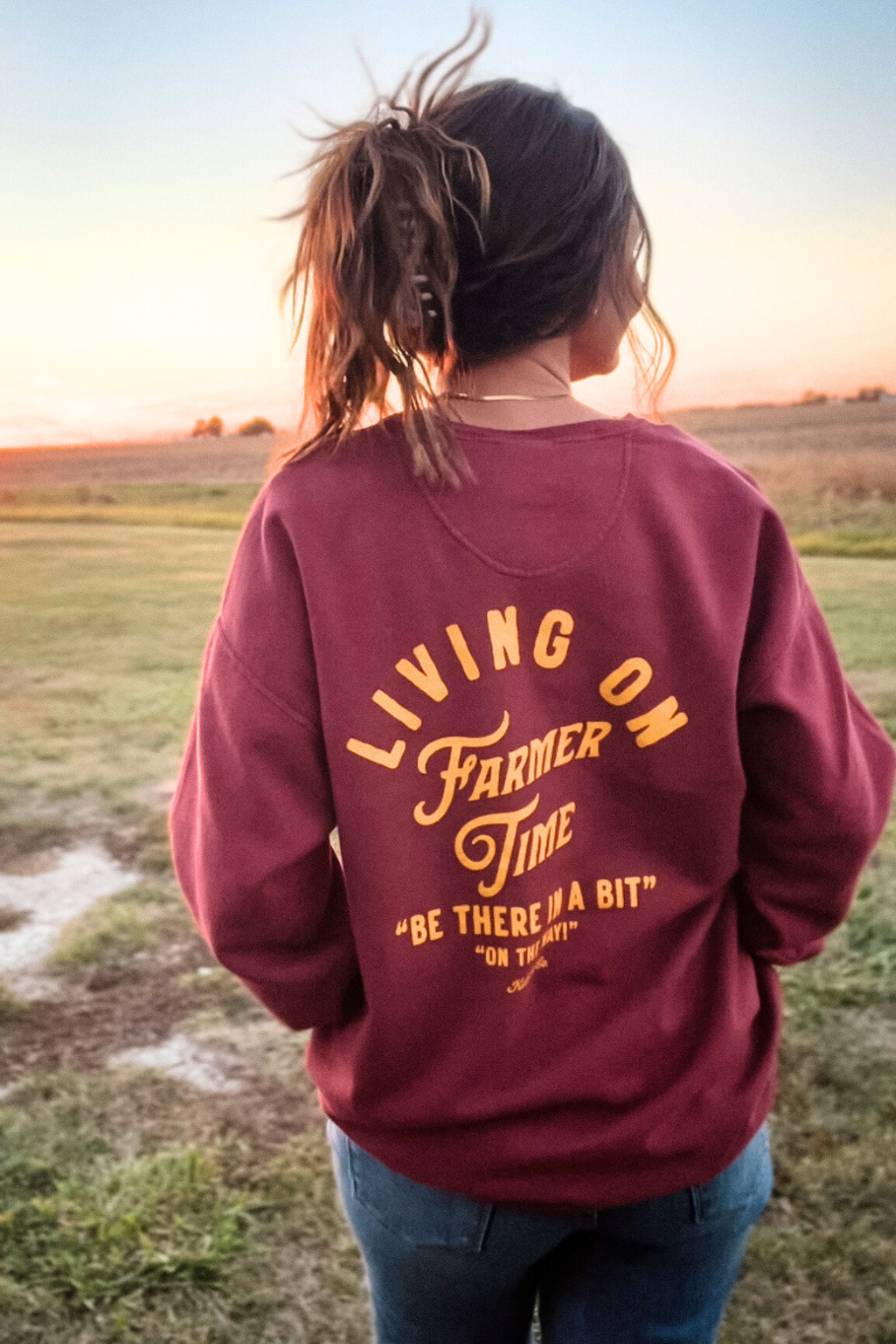 girl on farm standing in front of the sunset with the back of the sweatshirt saying living on farmer time