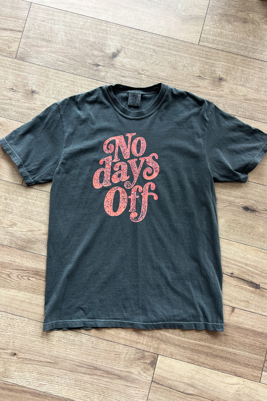 Flat lay of pepper gray t-shirt on a wooden floor, with coral text that read "No days off".