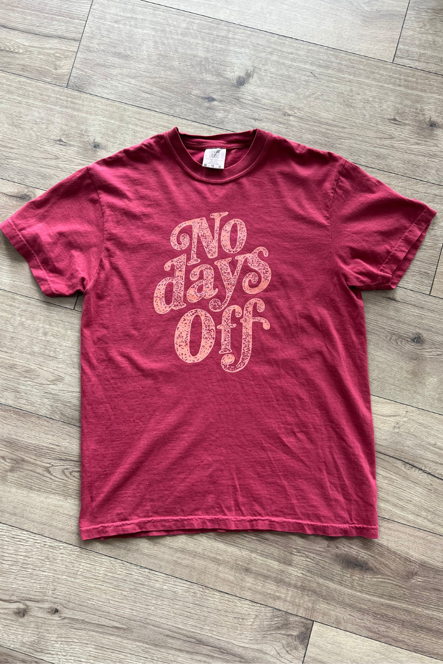 Flat lay of a tee in the color chili on a wooden floor, with text on the front of the shirt that reads "No days off" in coral. 