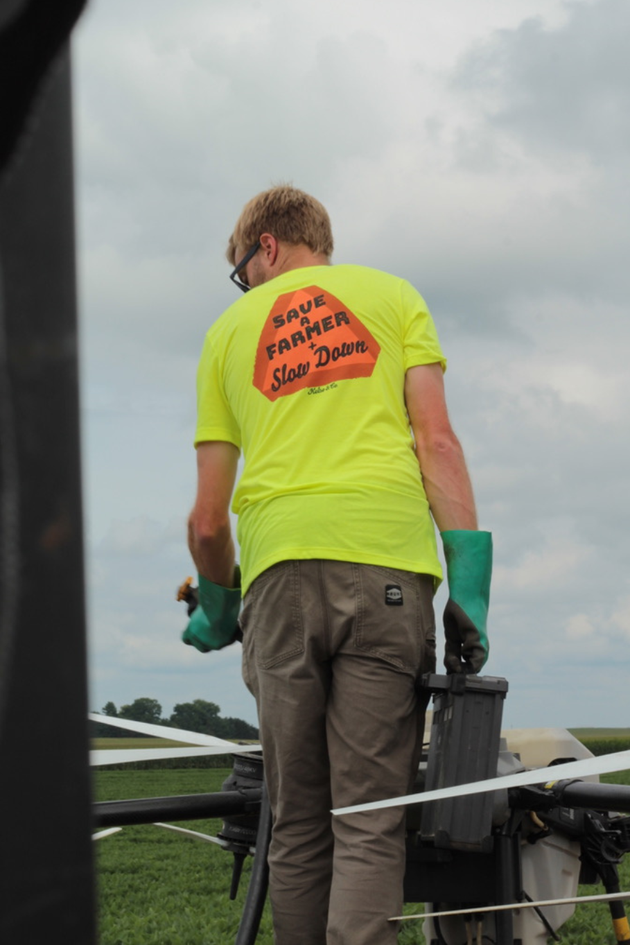Man wearing save a farmer slow down tshirt in neon yellow standing next to an agricultural drone