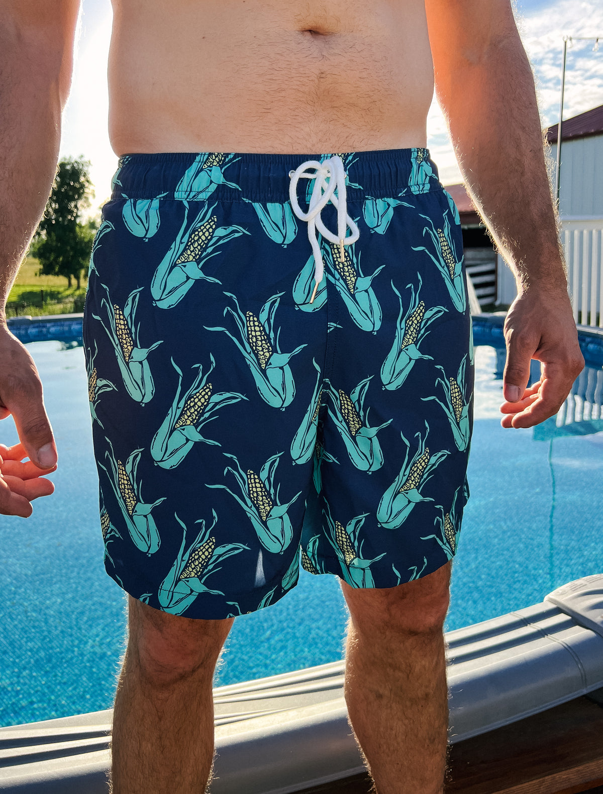Man standing in front of pool wearing the shuck yeah swim trunks that are navy blue  and have a corn print. 