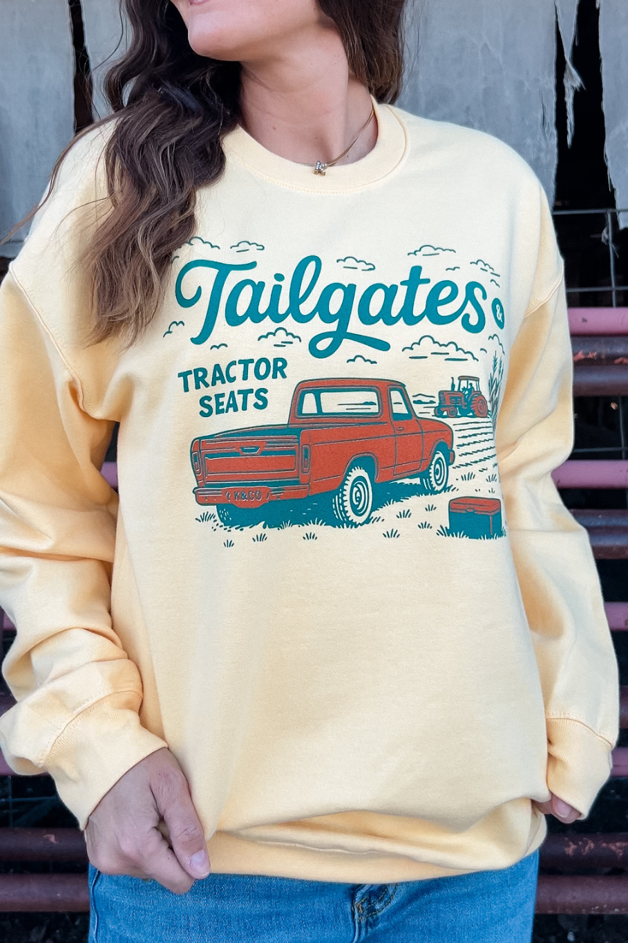 Girl wearing a light yellow sweatshirt with a graphic that has an old pick up truck and a tractor working a corn field that says &quot;tailgates &amp; Tractor seats&quot; in teal and orange