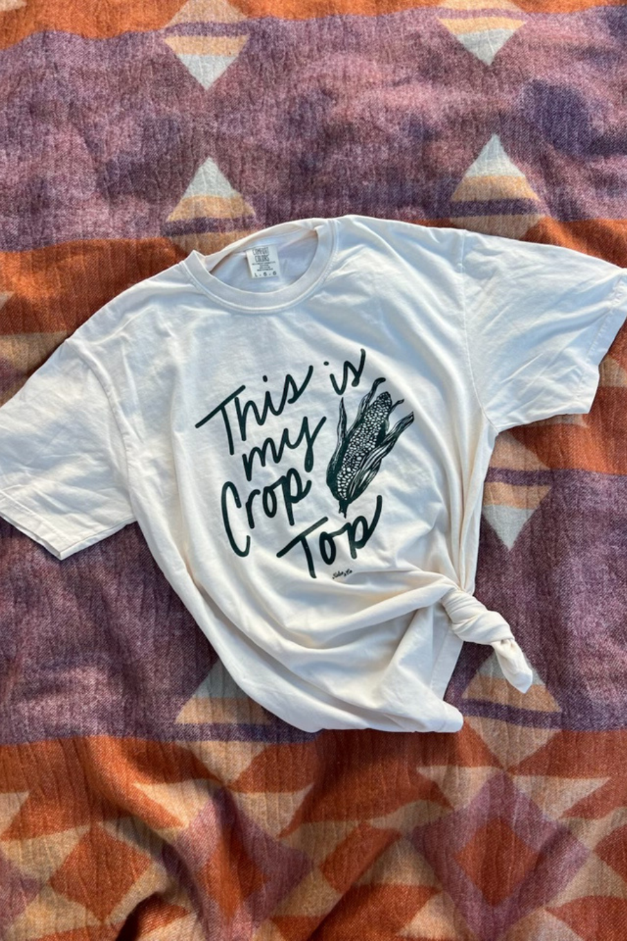 A ivory graphic tee with a knot at the hem, featuring the text 'This is my Crop Top' and a corn cob graphic, laid flat on a patterned blanket.