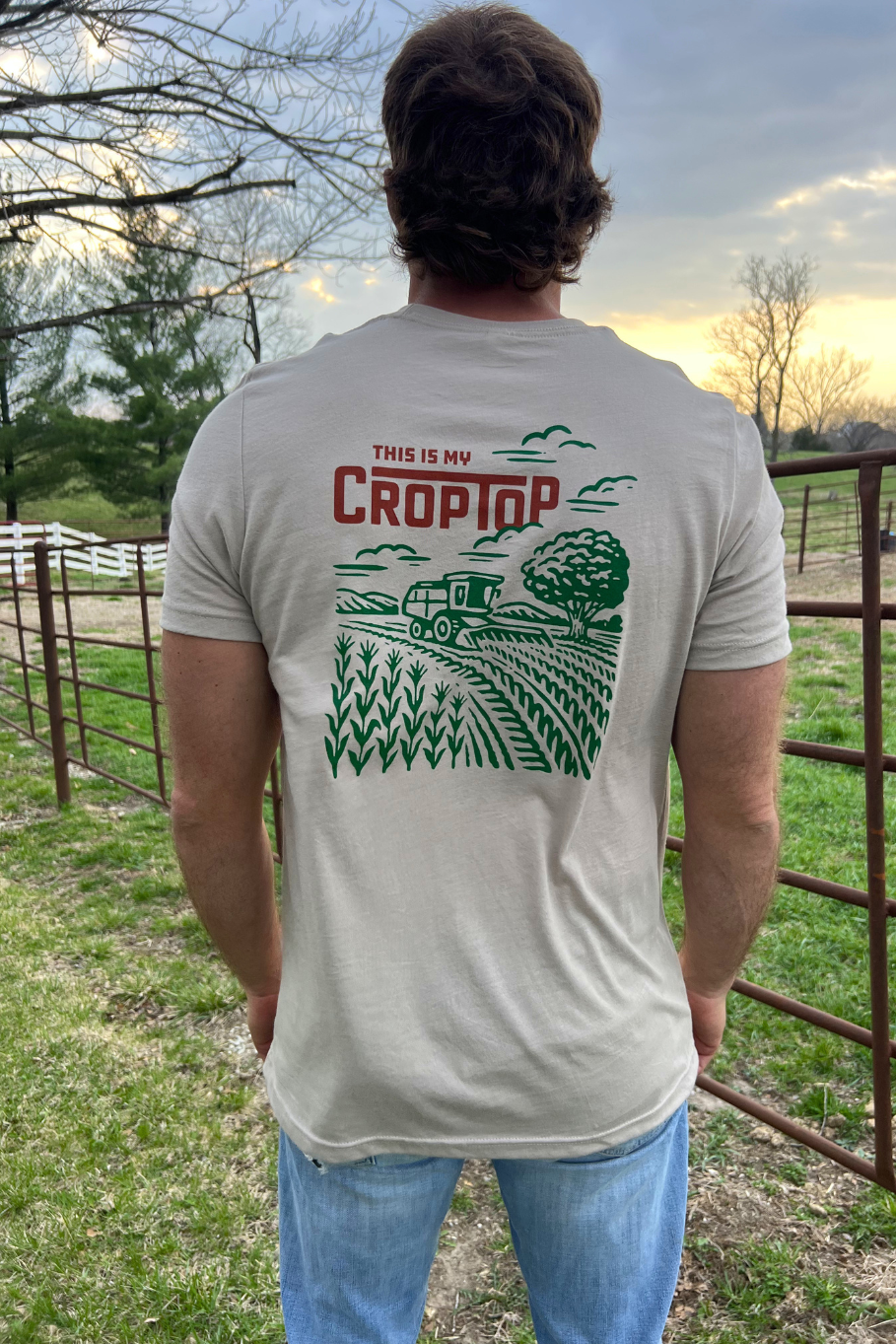 guy on a farm wearing a tan tshirt, the back of the tshirt says "this is my crop top" with a graphic of a combine in a corn field