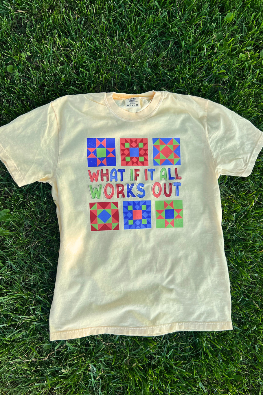 flatlay of a butter yellow comfort colors graphic tee on grass with a multi colored barn quilt design that says "what if it all works out" 