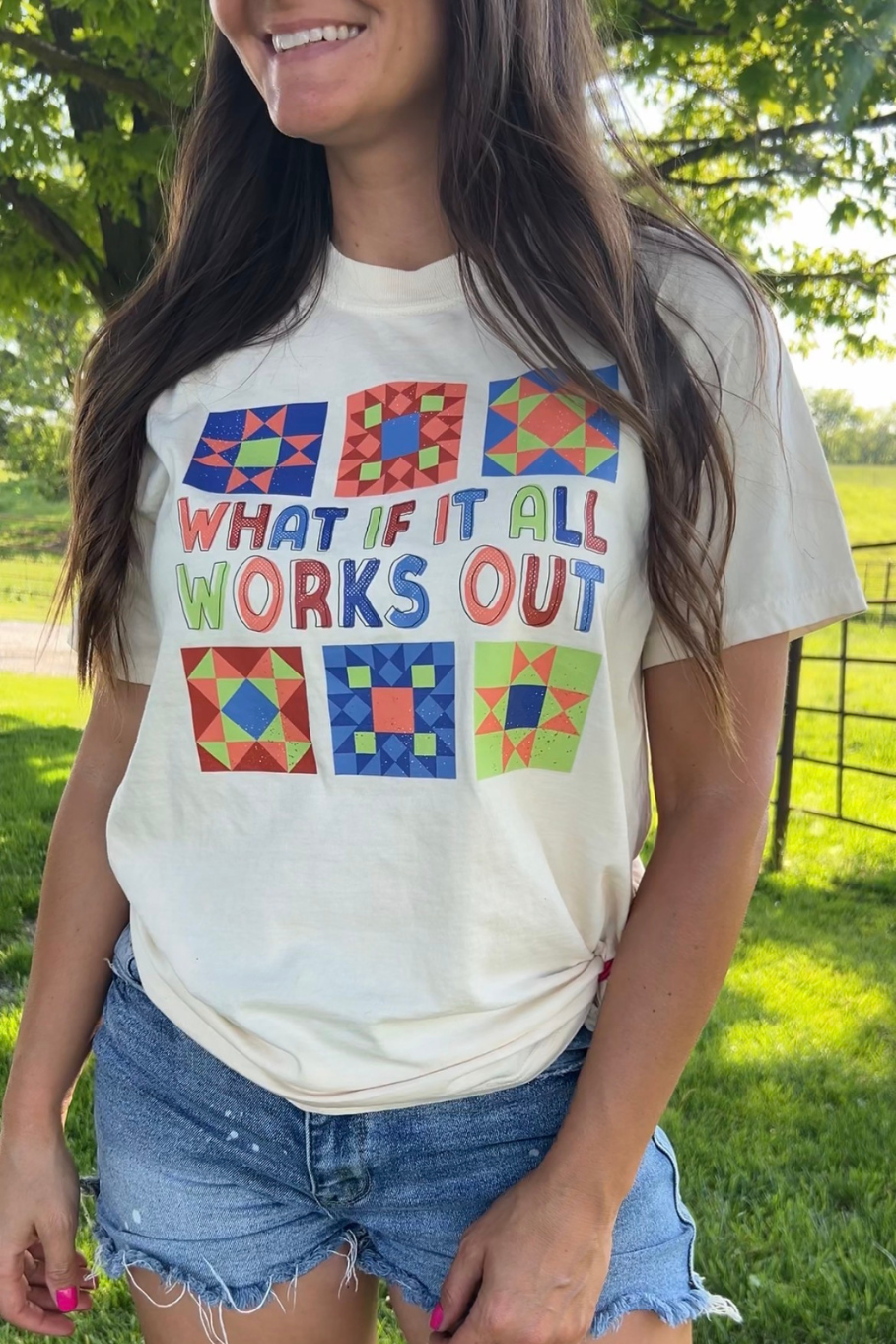 Woman wearing ivory tee with multicolored graphic featuring 6 barn quilt patterns and says "what if it all works out"