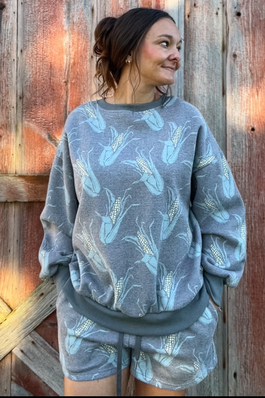 Woman standing in front of wooden barn door wearing a crewneck and sweat short set in a faded charcoal color with ears of corn scattered. 