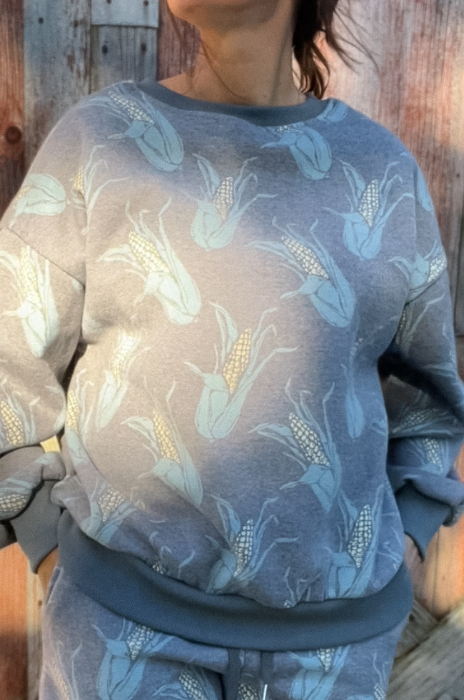 Woman standing in front of wooden barn door wearing a crewneck and sweat short set in a faded charcoal color with ears of corn scattered, with her hands in her pockets. 