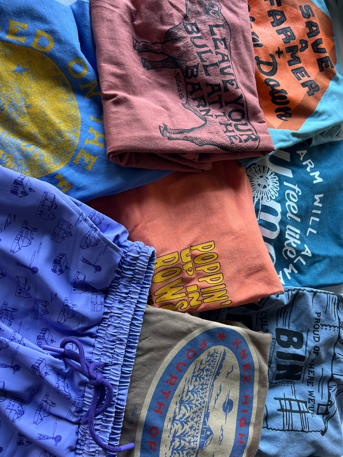 A pile of folded graphic t-shirts and shorts in assorted colors, including blue, pink, orange, purple, and gray, each featuring vintage-style printed graphics and text.
