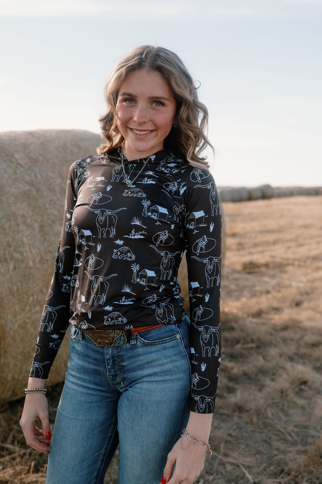 girl in front of a round hay bale wearing dark brown performance hoodie with light blue doodles of western scenes - armadillo, longhorn, barn with a windmill, cowboys, and desert brush