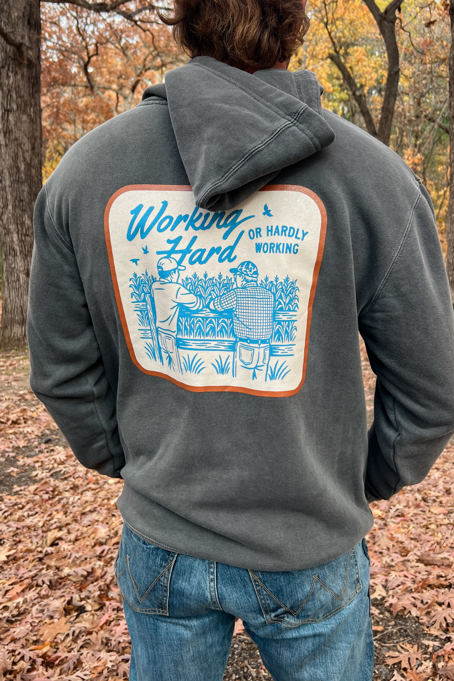 back of a guy wearing a pigmented black hoodie with working hard hardly working graphic - two farmers leaning against a fence staring out over a cornfield