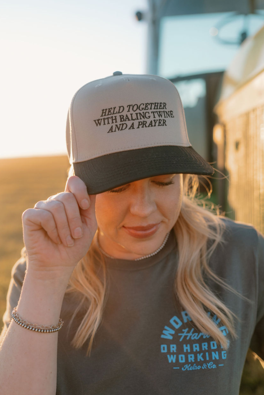 woman wearing a khaki trucker hat with a black bill that is embroidered "held together with baling twine and a prayer"