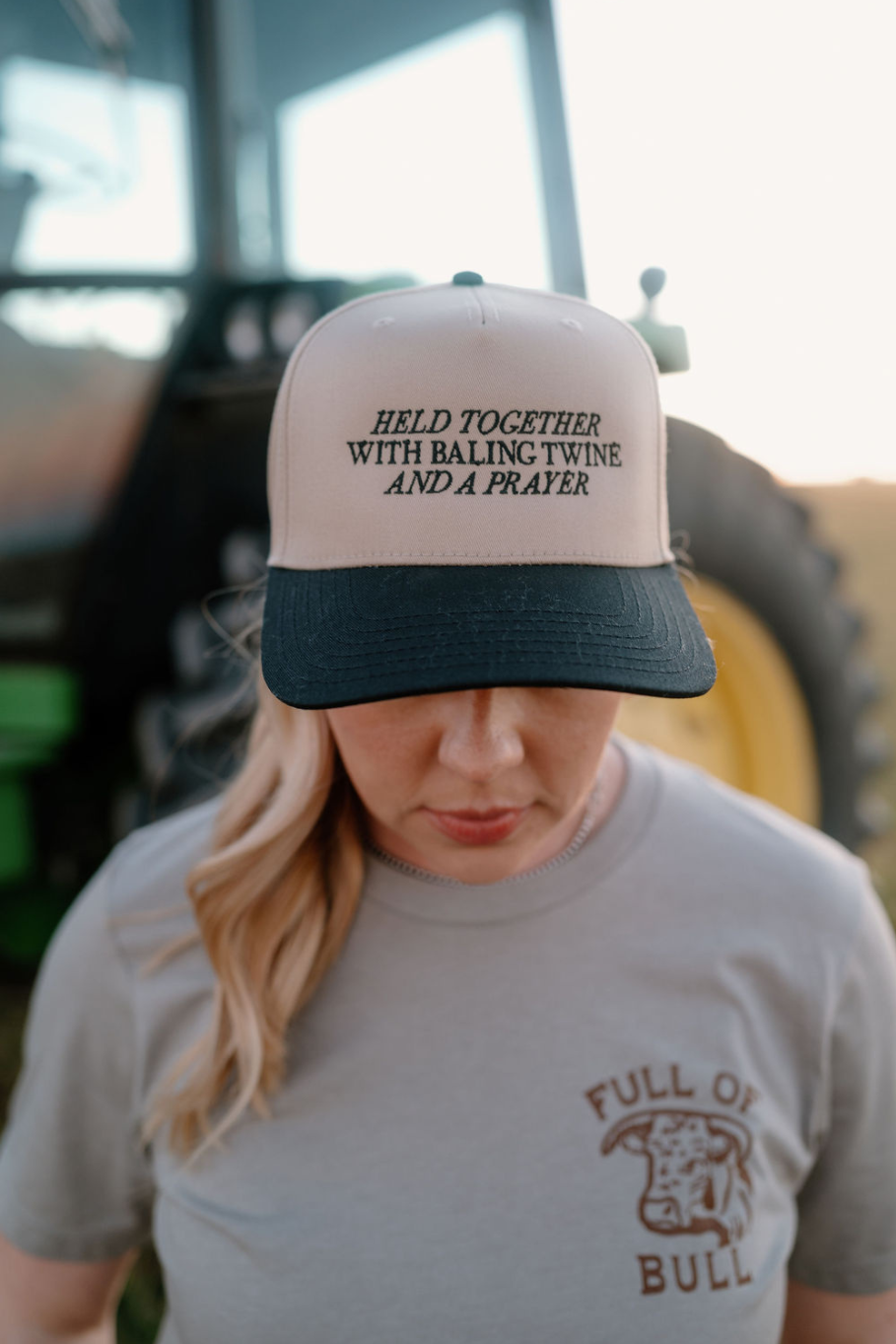 women standing in front of a tractor with a trucker hat on. Khaki hat with black bill that is embroidered "held together with baling twine and a prayer"