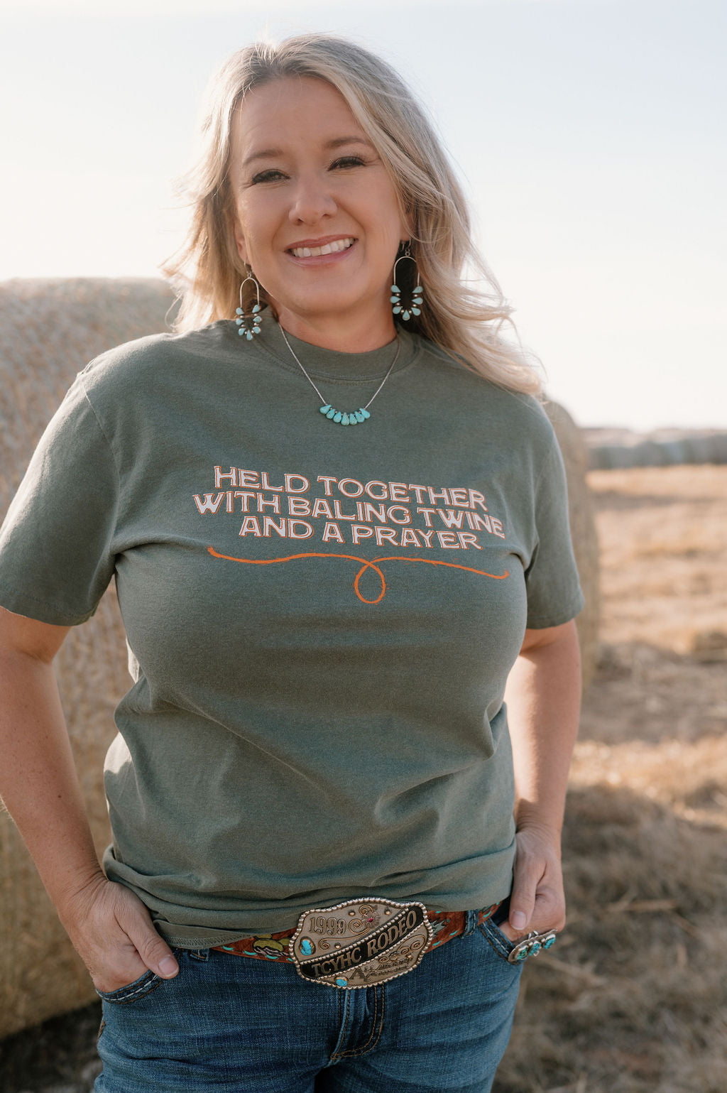 woman on a farm wearing a moss colored tshirt with cream and orange colored graphic that says "held together with baling twine and a prayer"