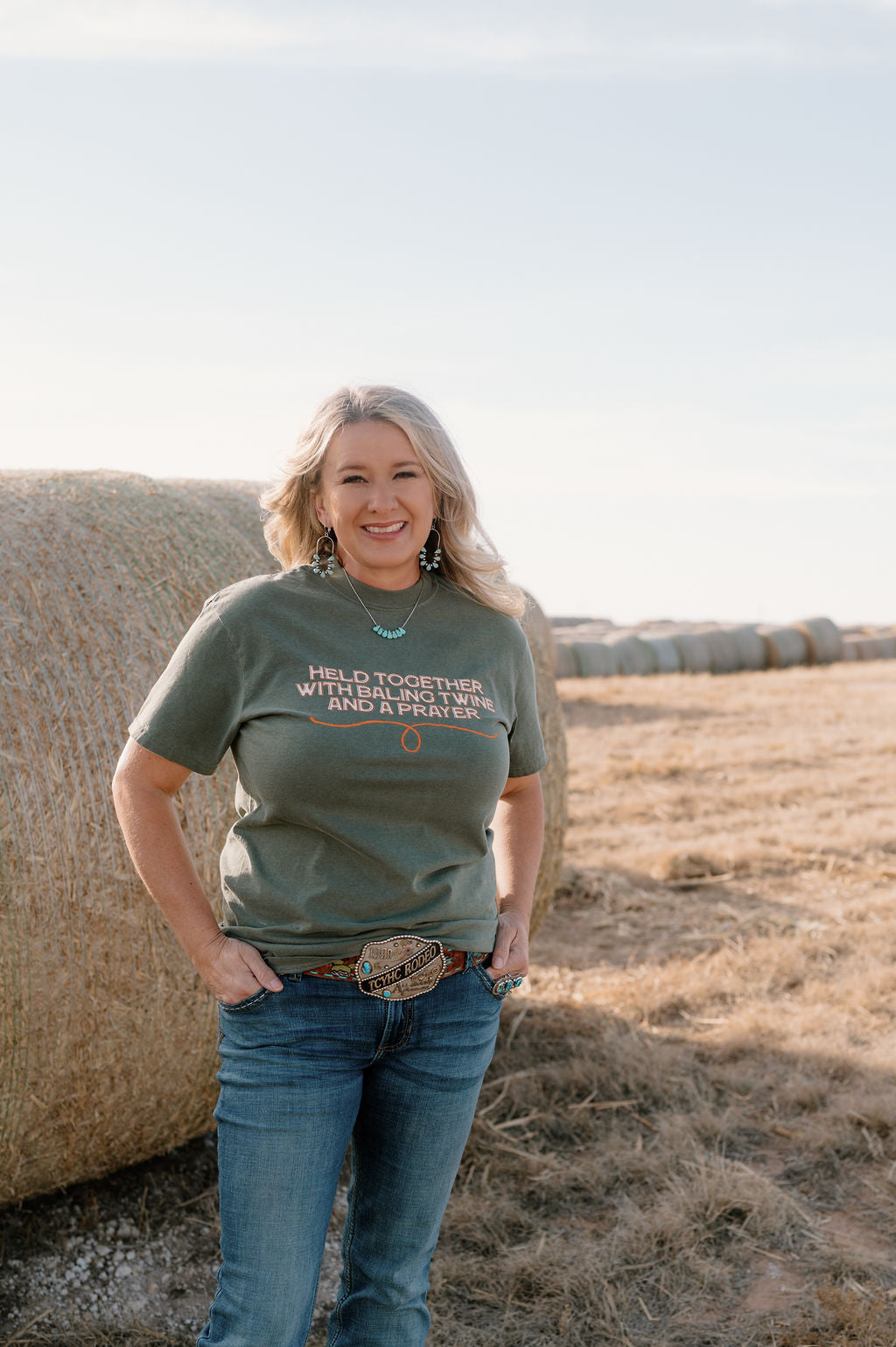 woman on a farm in a moss green colored tshirt with cream and orange text that says "held together with baling twine and a prayer"