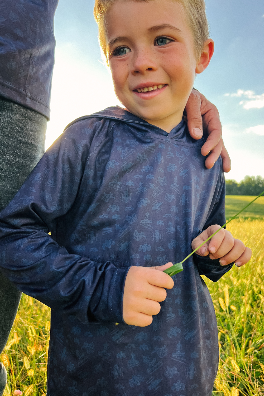 boy wearing blue performance hoodie with tractors on it in a field of grass