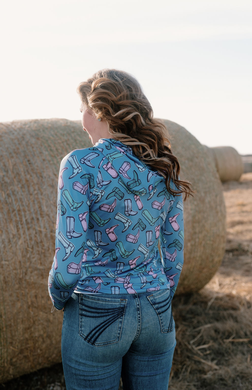 girl on a farm in front of hay bales with the boot scootin performance hoodie - a periwinkle base with multip colored muted cowgirl boots in greens, blues and purples