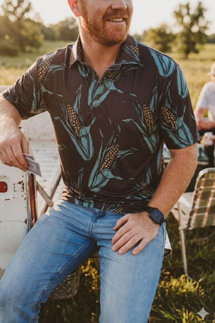 guy sitting on the tailgate of a truck playing cards wearing a brown kelso and co polo with large ears of corn all over it