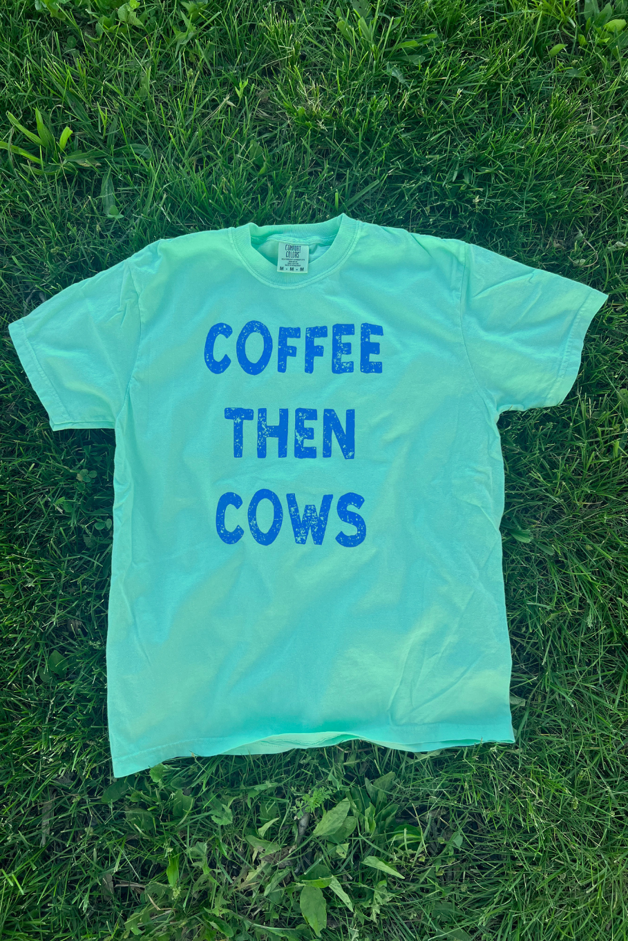 Teal t shirt with royal blue text "Coffee then cows" on front of the shirt, laying on grass. 
