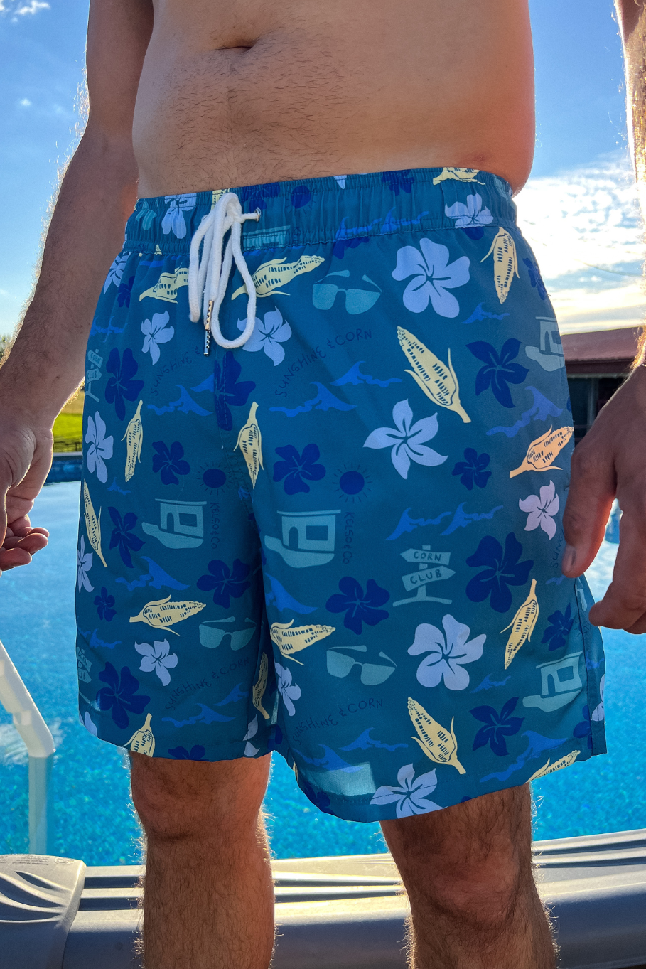 guy standing in from of an above ground pool with teal swim trunks on featuring a variety of summer motifs like hawaiian flowers, ears of corn, sun glasses, and a sign that says "corn club"