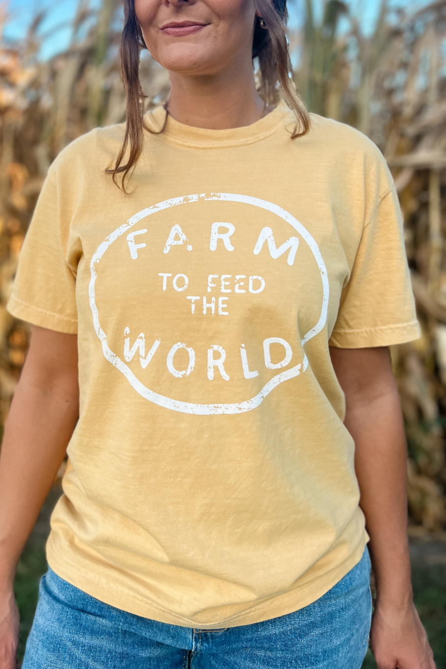 girl standing in front of a corn field with a comfort colors mustard tshirt on that says farm to feed the world