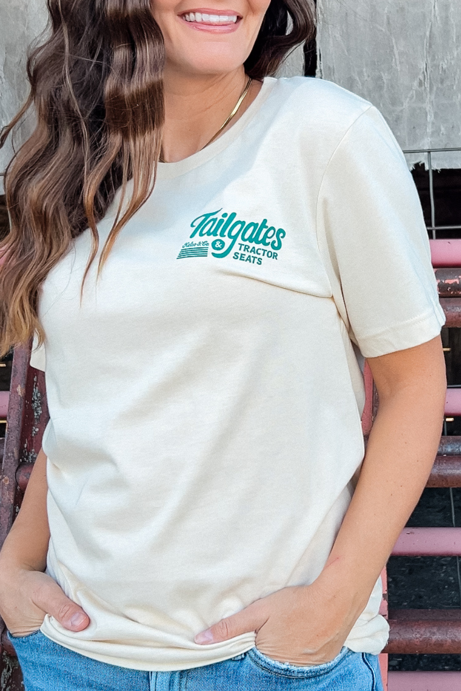 girl standing in front of cattle panels with her hand in her pocket, showing the front of her t-shirt that hsays &quot;tailgates &amp; Tractor seats&quot; on the left chest in teal