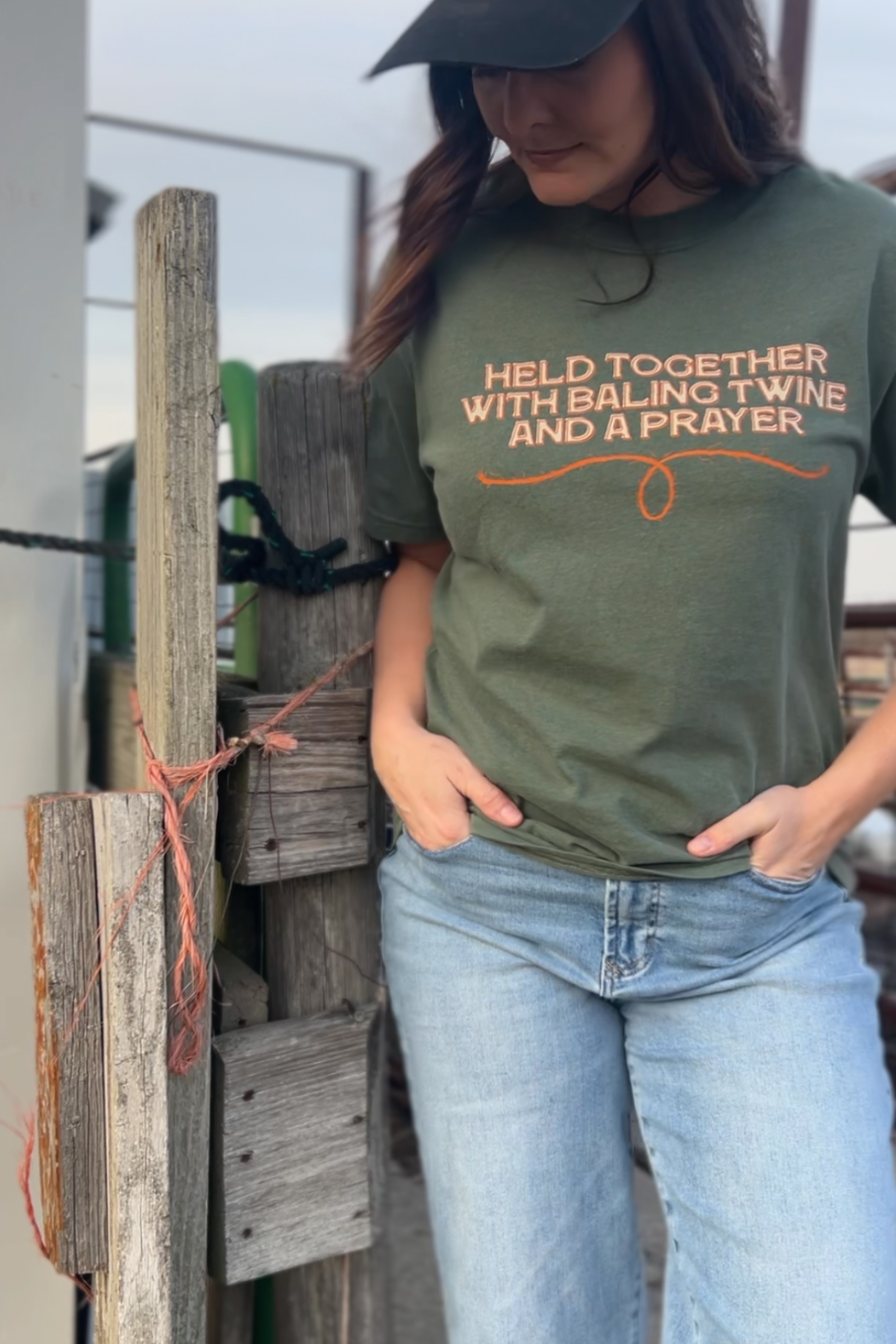 girl leaning up against a fence with a moss t-shirt on that says held together with baling twine and a prayer 