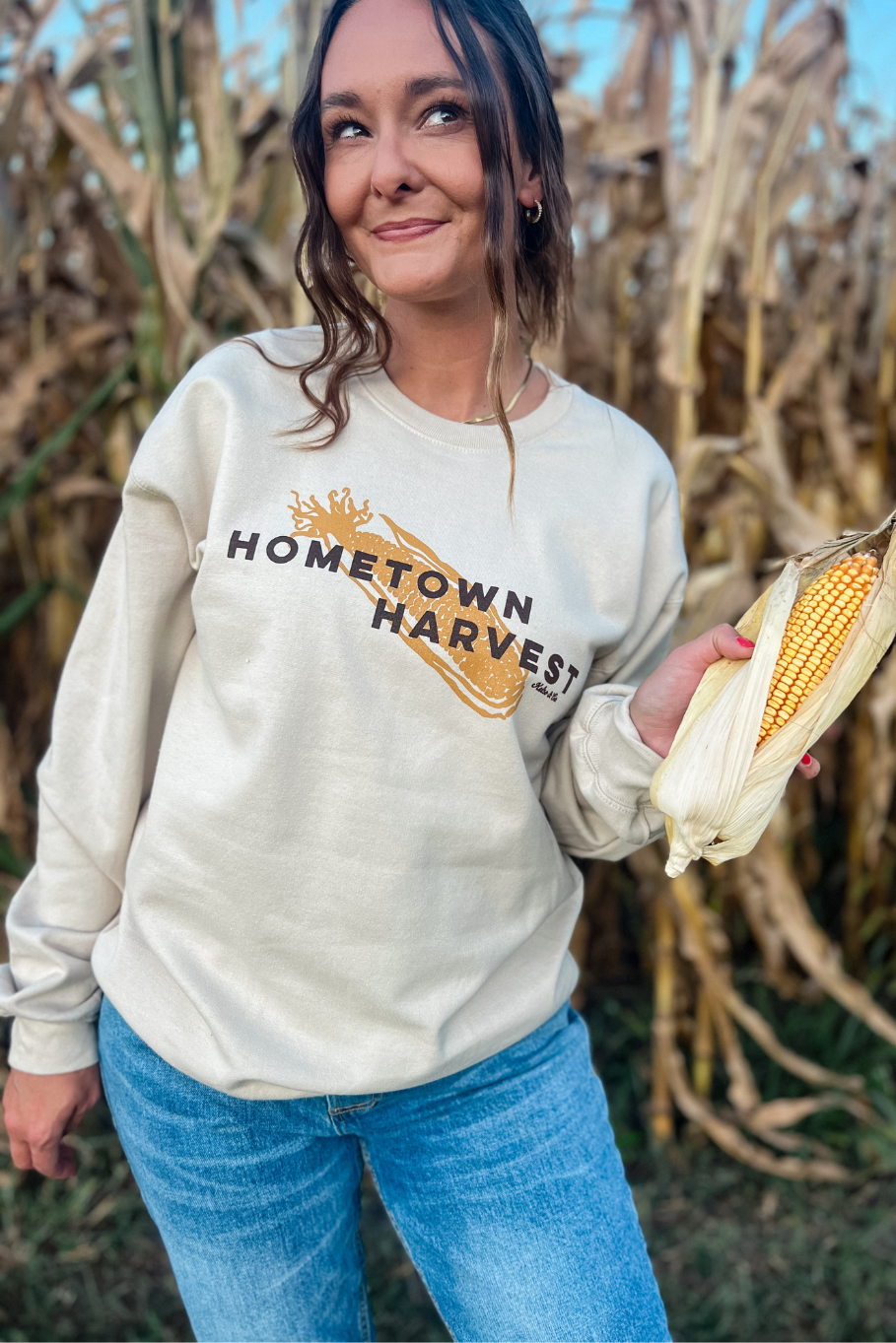 girl wearing hometown harvest crewneck in with an ear of corn in front of a corn field