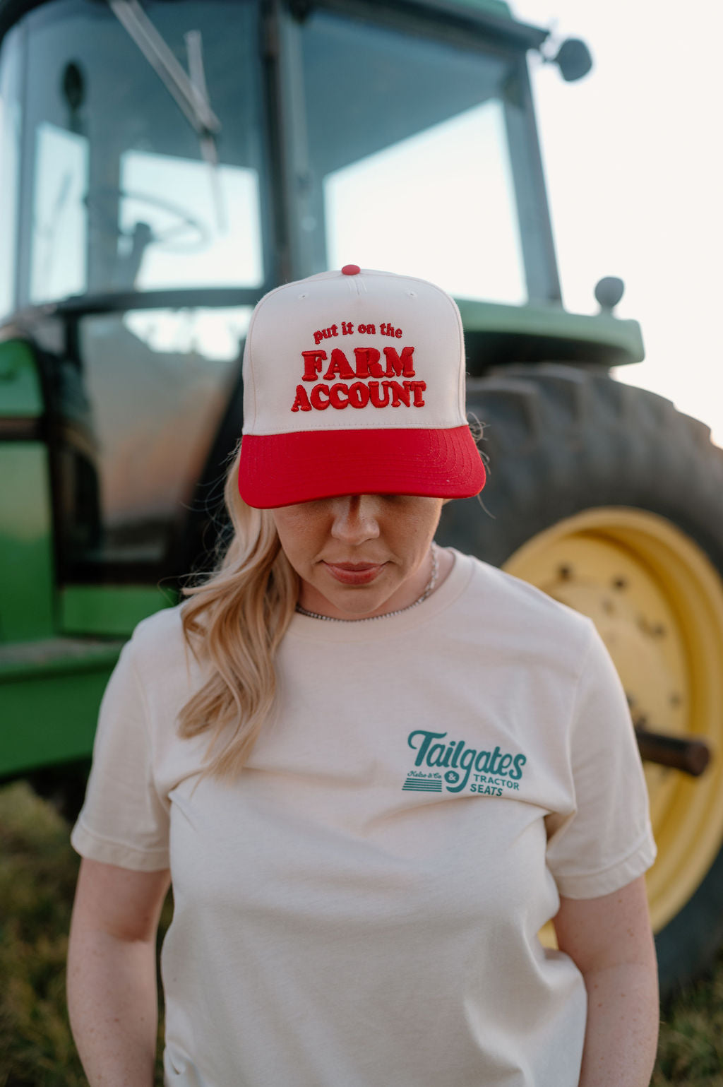 Red and natural trucker hat on girl standing in front of a tractor. Hat is embroidered with "Put it on the farm account"in red