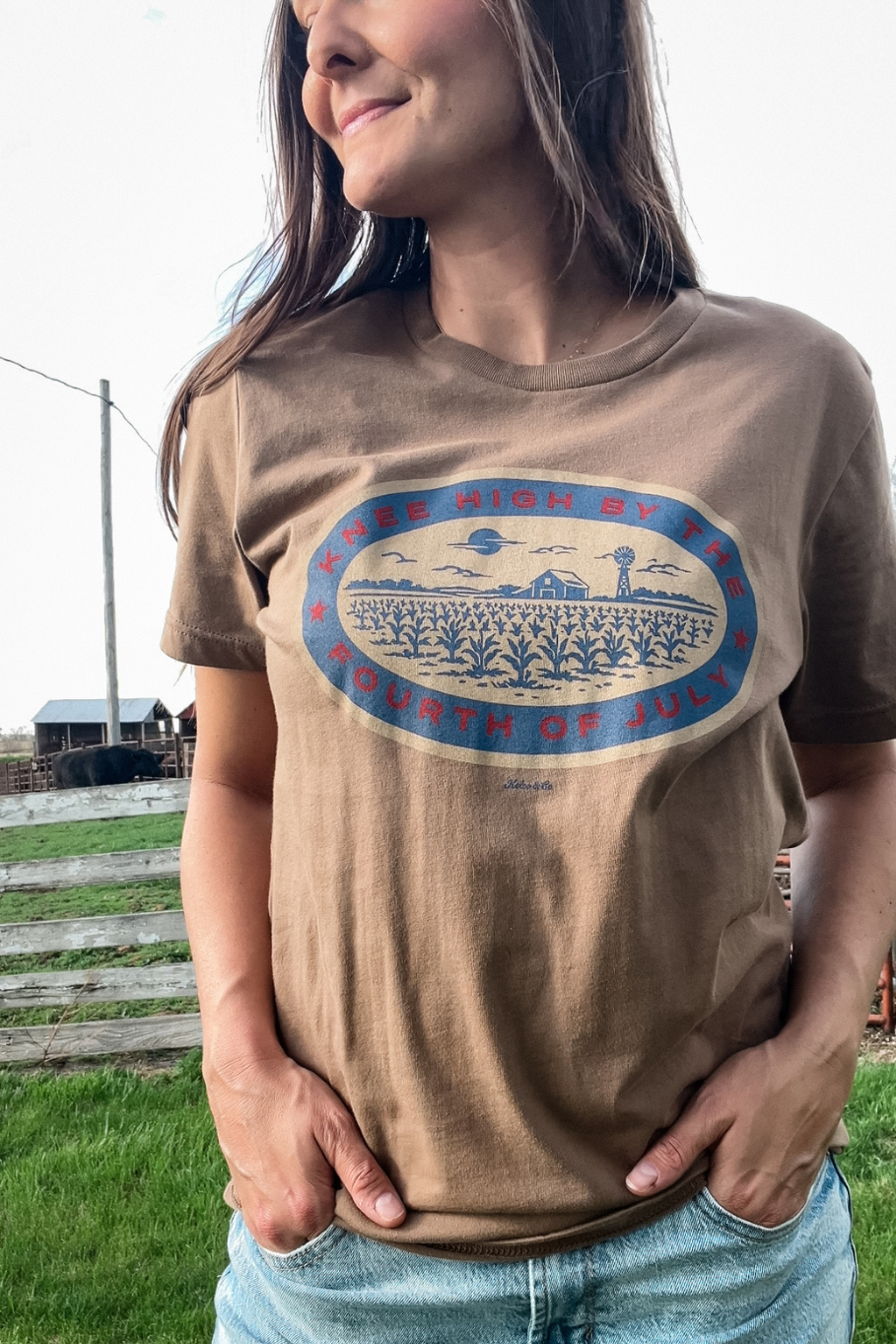 woman in front of a white fence on a farm with a light brown tshirt on and a graphic in blue, red and tan that says "knee high by the fourth of july." The graphic shows a corn field with a barn and windmill