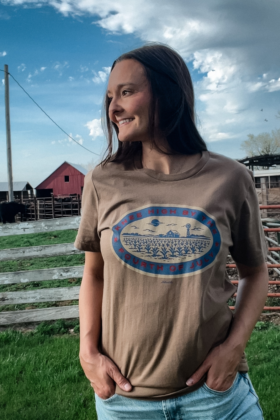 woman in front of a white fence on a farm with a light brown tshirt on and a graphic in blue, red and tan that says "knee high by the fourth of july." The graphic shows a corn field with a barn and windmill