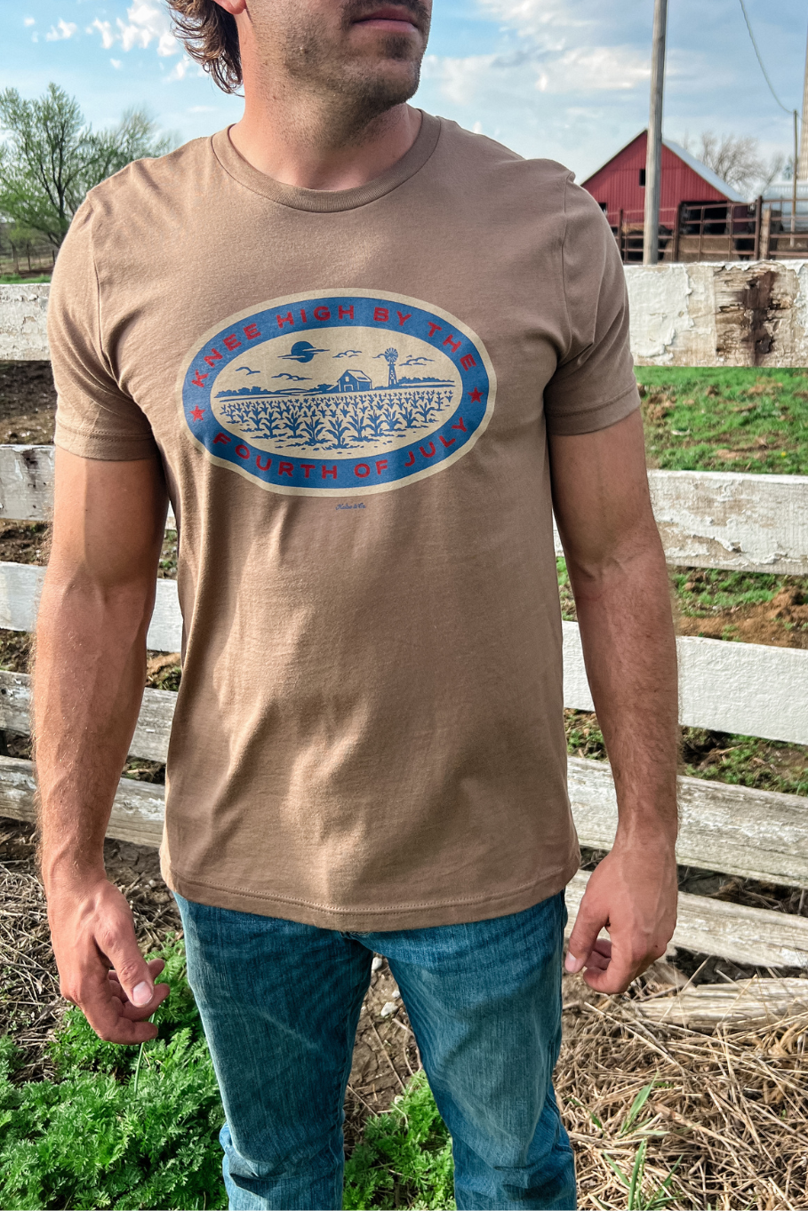 Guy in front of a white fence on a farm with a light brown tshirt on and a graphic in blue, red and tan that says "knee high by the fourth of july." The graphic shows a corn field with a barn and windmill