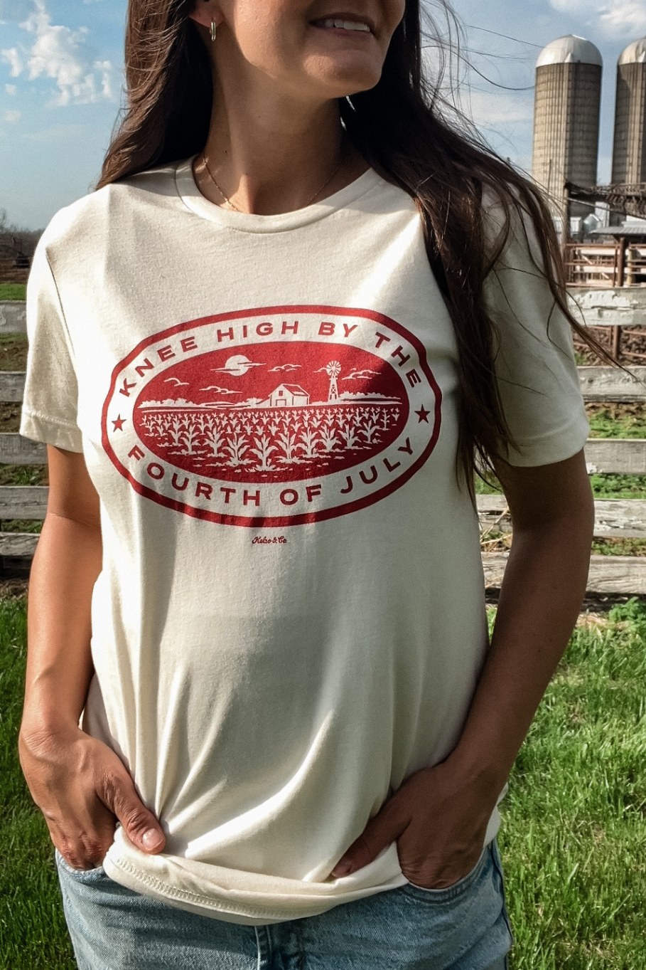 woman standing on a farm in a natural colored graphic tee with a graph graphic that says knee high by the fourth of july