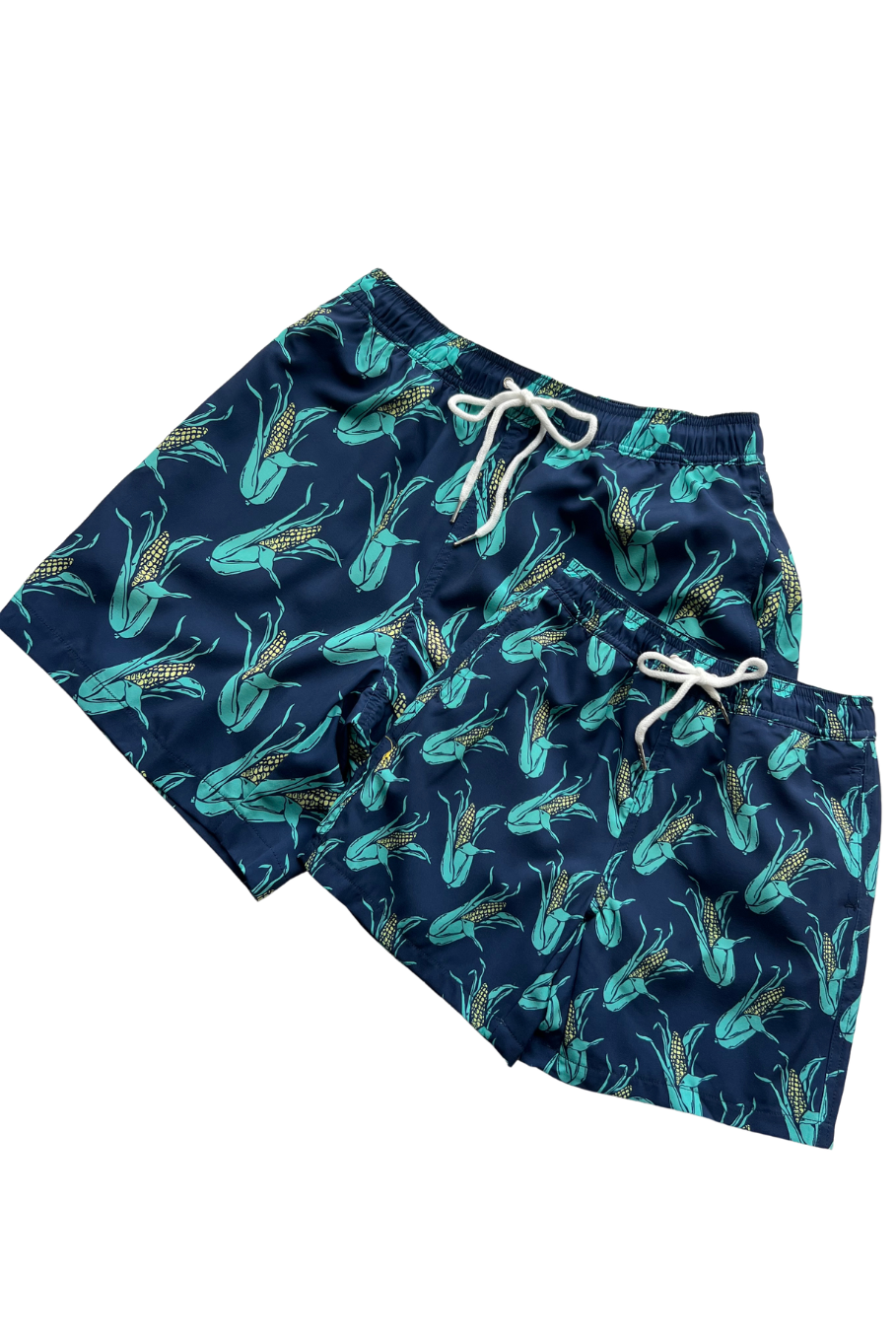 Flat lay of of boy's navy blue swim trunks with teal and yellow ears of corn over the matching men's trunks. 