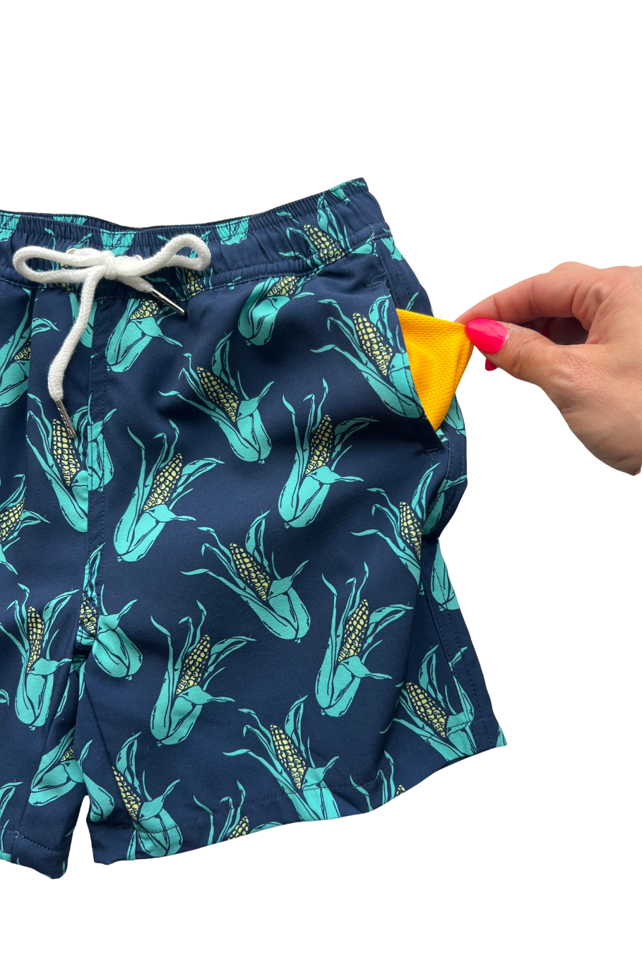 Flat lay of boy's navy blue swim trunks with teal and yellow ears of corn with a woman's hand pulling out the mesh lining in the pocket. 