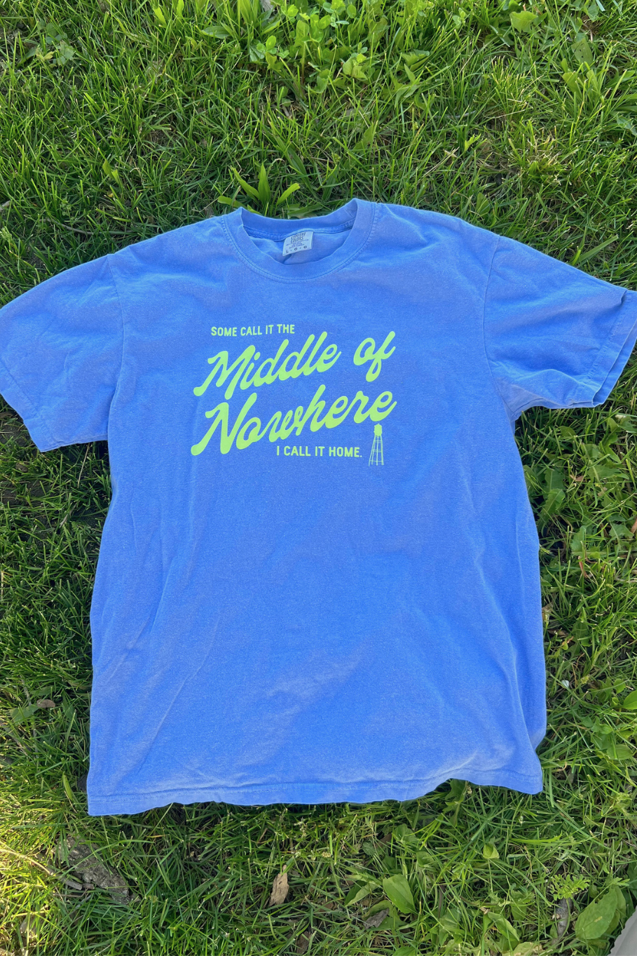 flo blue comfort colors tee laying on grass with a lime green graphic "some call it the middle of nowhere I call it home" with a lime green graphic of a water tower