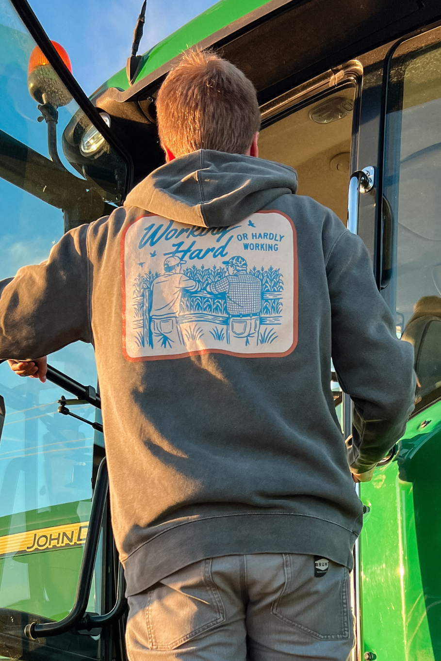Guy climbing into a John Deere tractor wearing a farm luxury hoodie that shows two farmers leaning up against a fence talking and says &quot;working hard or hardly working&quot;