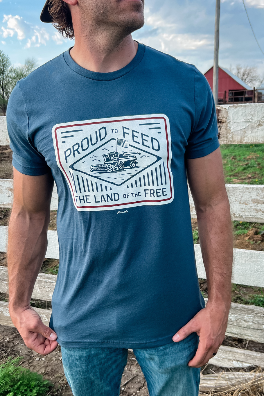guy in front of a white fence with a blue graphic tshirt on that says "proud to feed the land of the free." The graphic has a combine with an american flag