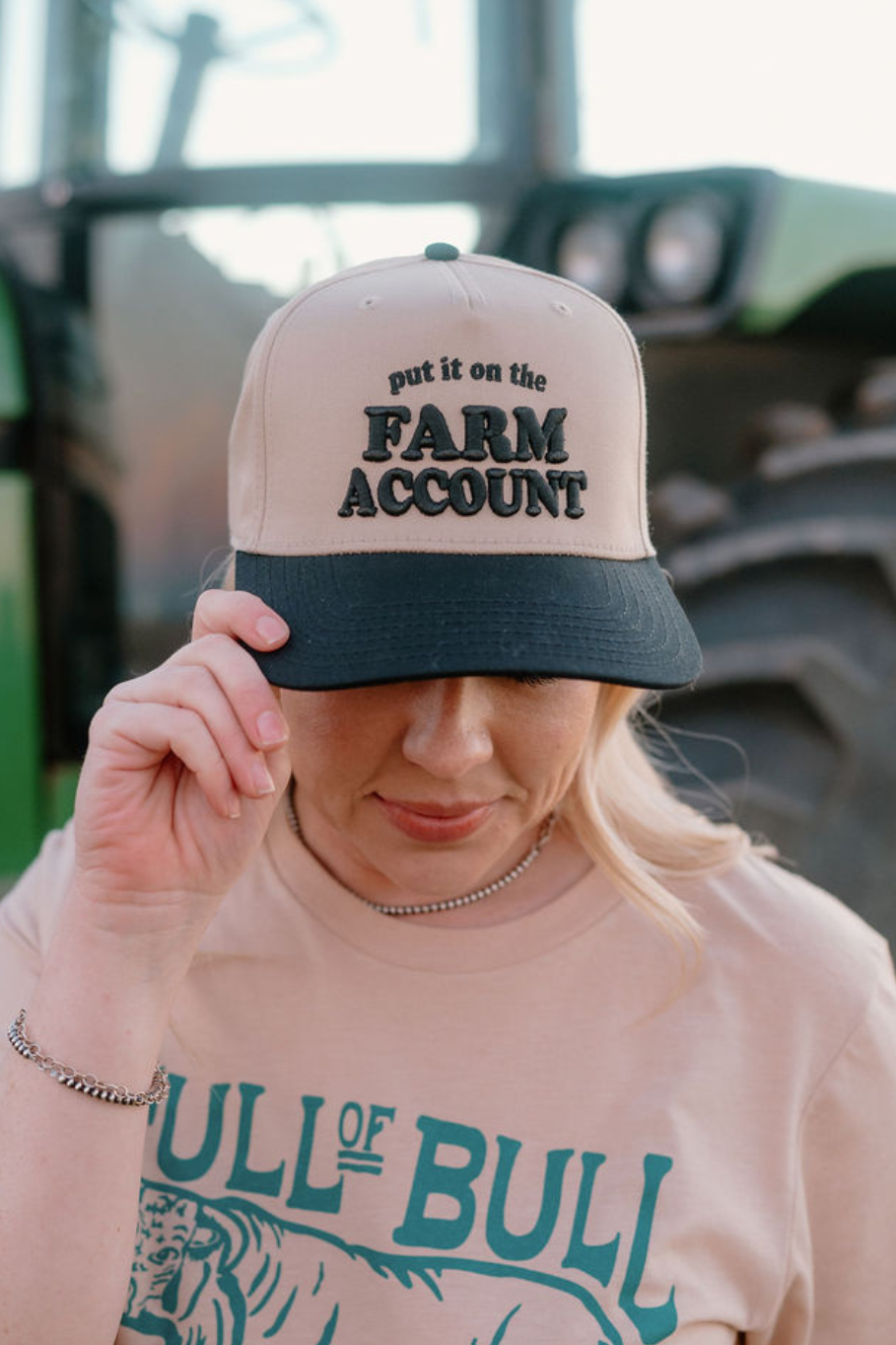 girl wearing a khaki trucker hat with a black bill that says "put it on the farm account" in embroidery