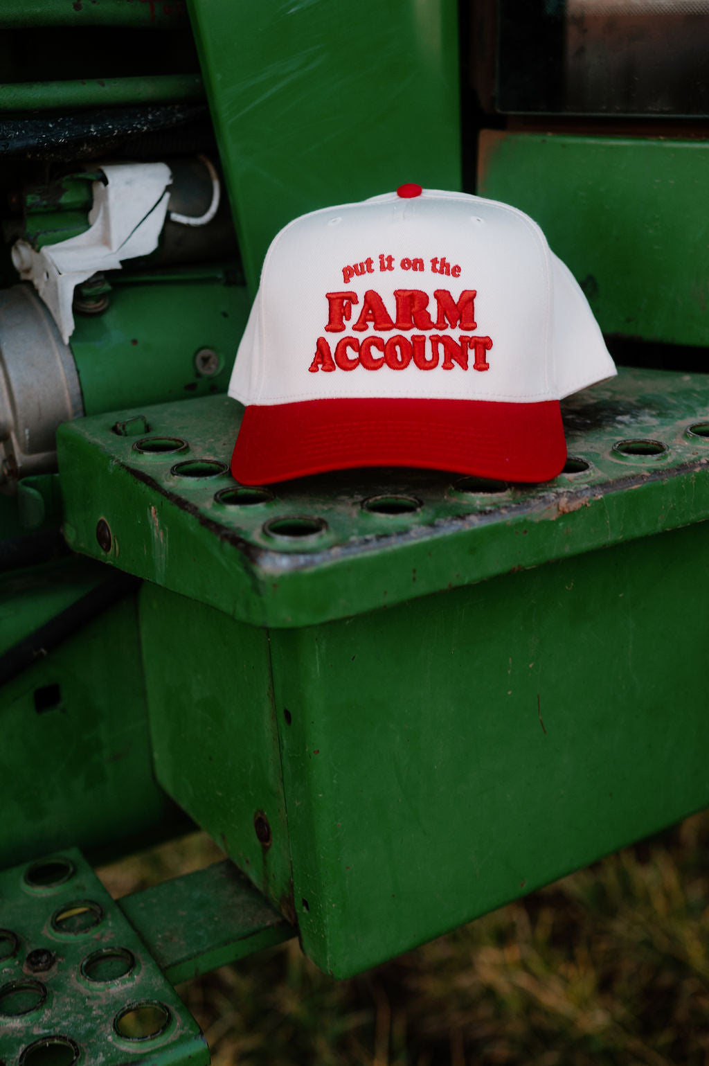 Red and natural trucker hat sitting on the step of a tractor. Hat is embroidered with "Put it on the farm account"in red