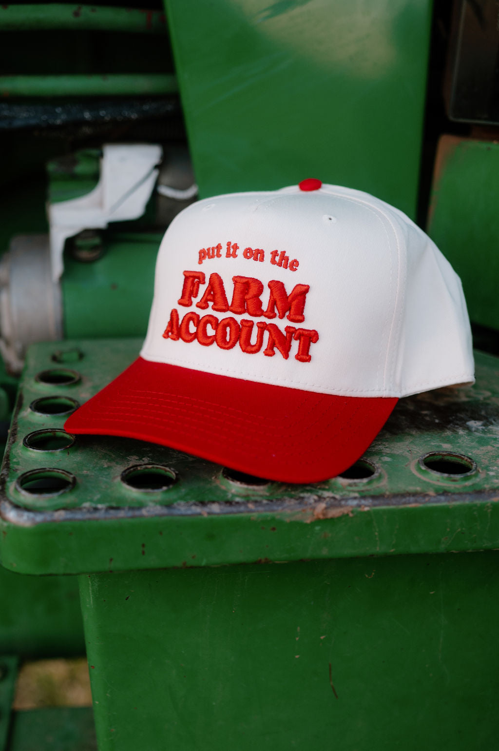Red and natural trucker hat sitting on the step of a tractor. Hat is embroidered with "Put it on the farm account"in red