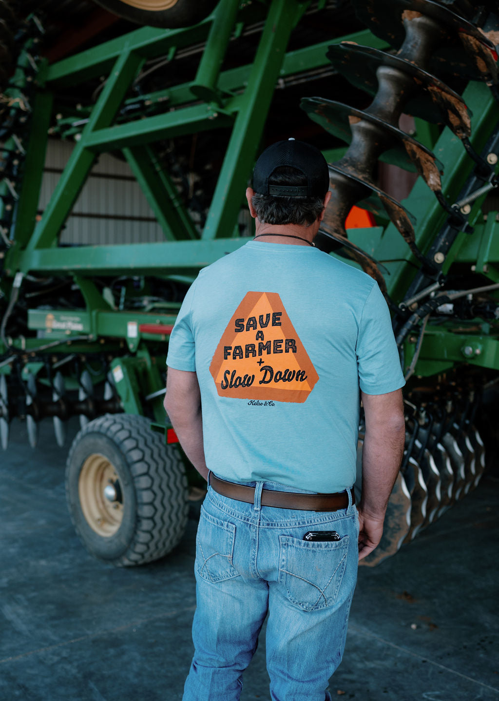 man wearing a blue tshirt in front of a farm implement, the back has an orange slow moving vehicle sign and says "save a farmer + slow down"