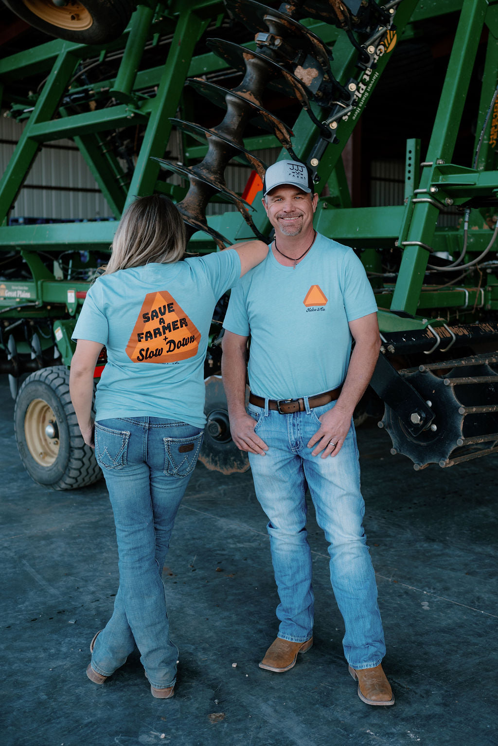 farm couple standing in front of a farm implement wearing a blue tshirt , the back has an orange slow moving vehicle sign and says "save a farmer + slow down," the front has a slow moving vehicle sign and the kelso & co logo
