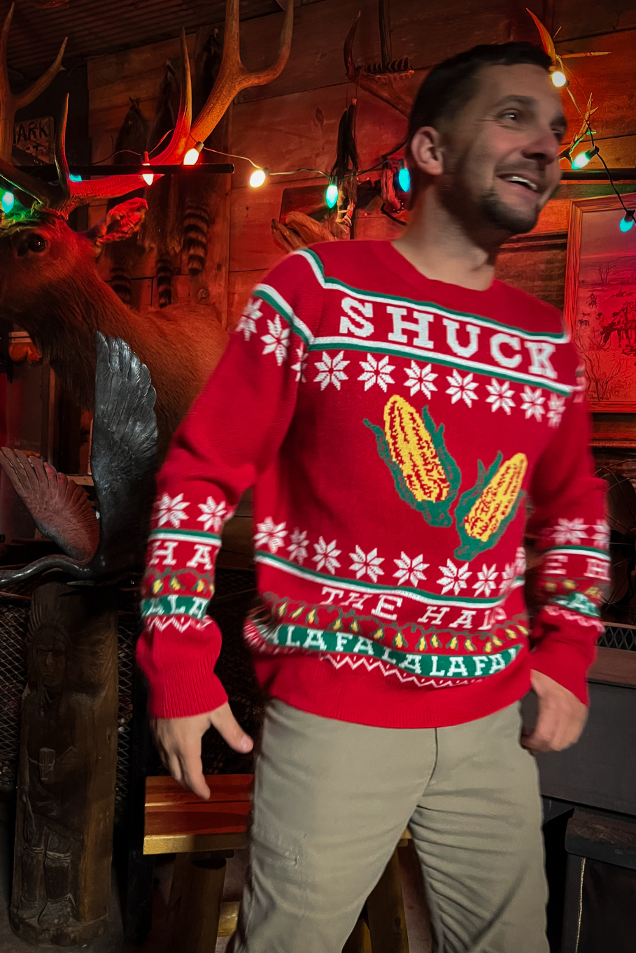 guy wearing red holiday sweater with ears of corn that says shuck the halls with colored christmas lights and an elk on the wall