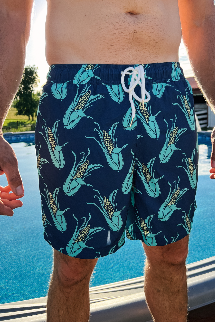man standing in front of an above ground pool with navy swim trunks on with ears of corn all over