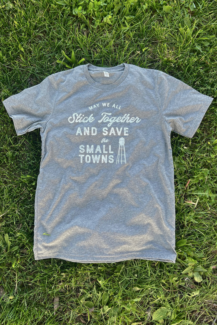 gray trebled tshirt laying on grass that says "may we all stick together and save the small towns" in white