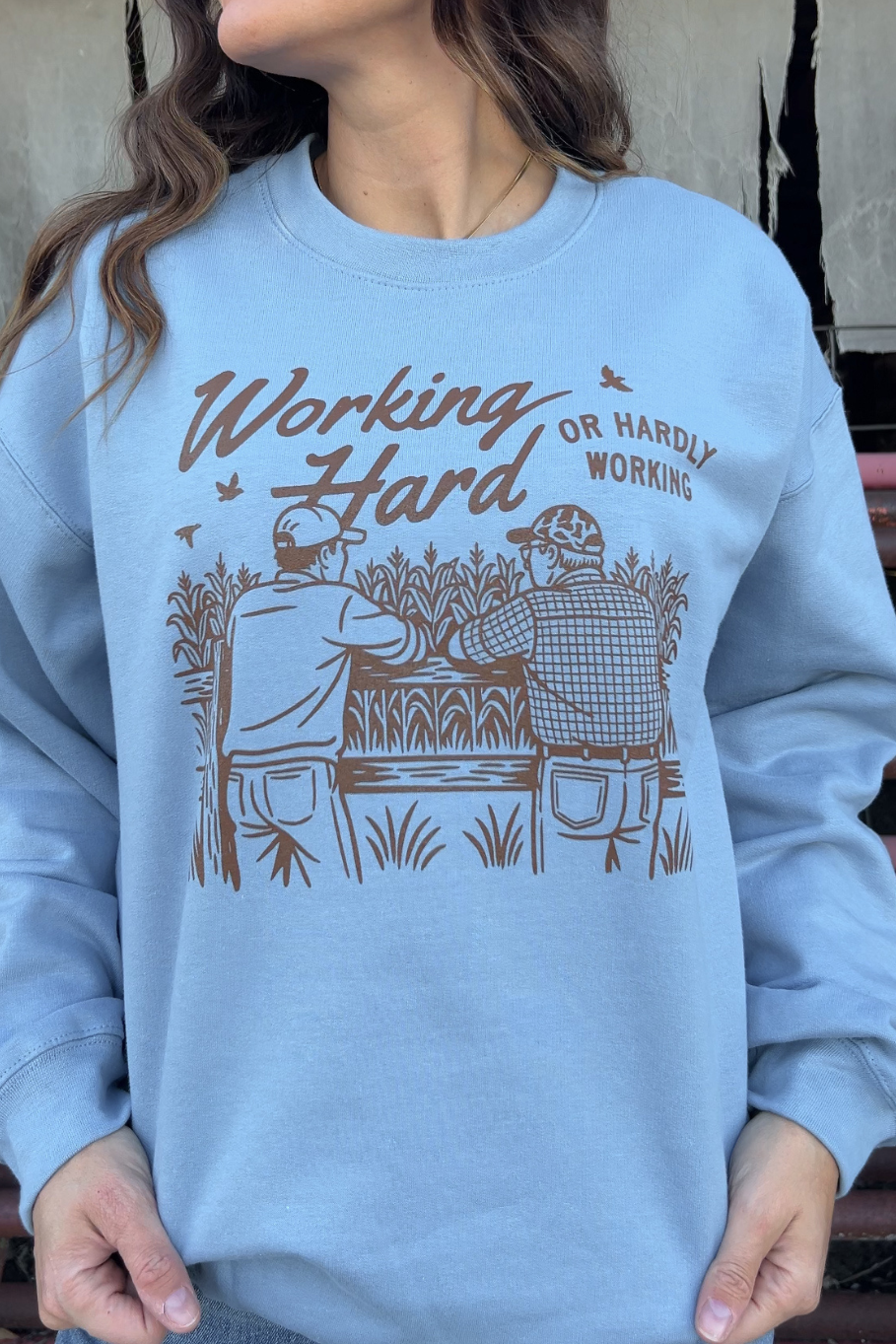 woman wearing a stone blue crewneck that has a graphic of two farmers leaning against a fence that says working hard or hardly working