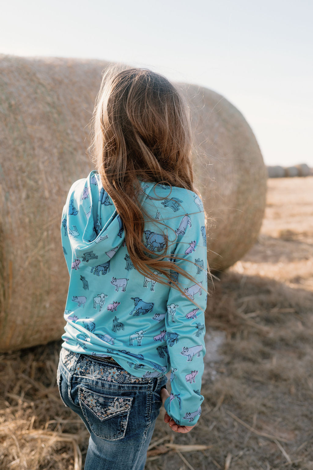 girl on a farm wearing a sea green performance hoodie with multicolored cows and pigs all over