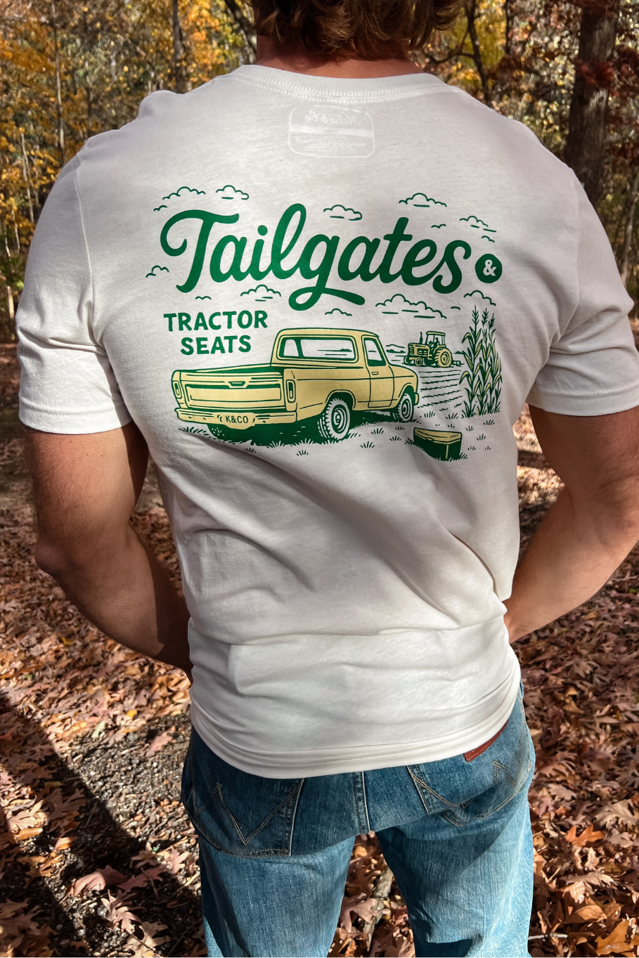 back of guy wearing vintage white graphic tee with tailgates and tractor seats graphic tee in green and gold with a graphic of a vintage truck and a tractor in a field