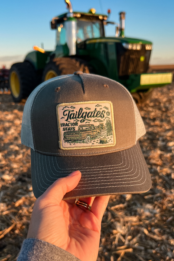 gray green richardson hat with embroidered patch that says tailgates and tractor seats in front of a john deere tractor in a corn field post harvest